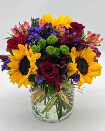 Reminiscent of strolling through a field of flowers, this colorful arrangement captures features magnificent sunflowers, alstroemeria and poms set in a trendy mason jar with a natural raffia bow.
